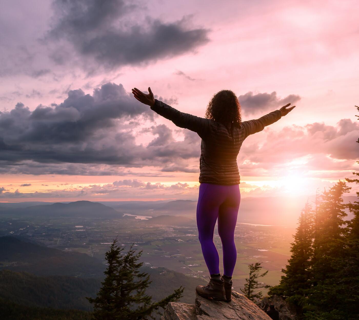 Woman standing on edge of a cliff as the sun rises, her arms are spread out in welcoming of the new day.